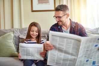 Vater sitzt mit kleiner Tochter auf dem Sofa beim gemeinsamen Zeitunglesen