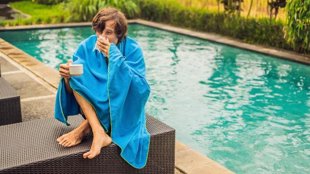
		Ein junger Mann sitzt am Pool mit einer Decke umschlungen und putzt sich die Nase
	