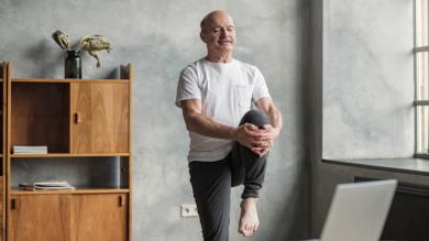 
		Ein älterer Man steht vor einem Laptop in seinem Wohnzimmer und macht eine Yoga-Übung, wo er auf einem Bein steht
	
