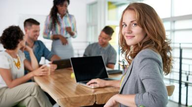 
		EIne Vereinbarkeitsmanager sitzt an einem Tisch mit ihrem Laptop und lacht in die Kamera. Ihre Kollegen sind verschwommen im Hintergrund zu sehen.
	