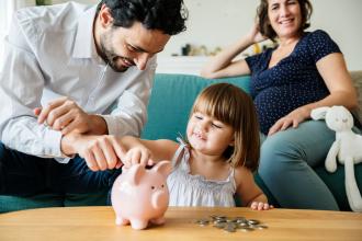 
		Ein schwangere Mutter sitzt mit ihrem Freund und ihrer Tochter im Wohnzimmer. Der Vater steckt mit der Tochter gemeinsam eine Geldmünze in ein Sparschwein auf dem Couchtisch.
	