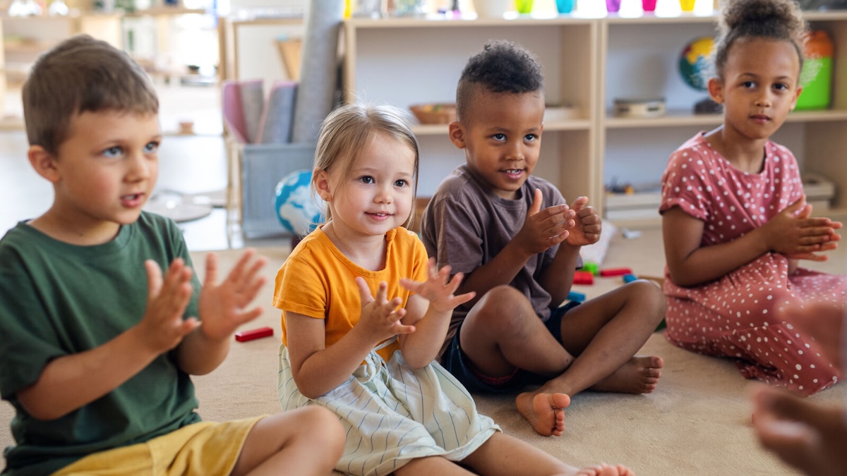Vier Kleinkinder sitzen nebeneinander, klatschen und schauen zur Betreuungsperson