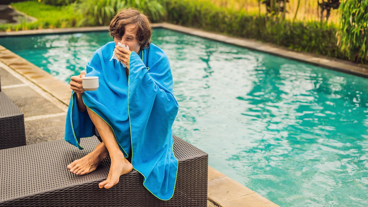 Ein junger Mann sitzt am Pool mit einer Decke umschlungen und putzt sich die Nase